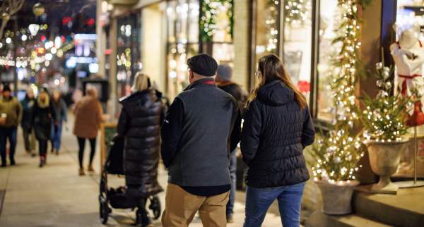 People walk along a city sidewalk decorated with holiday lights and storefront displays at night