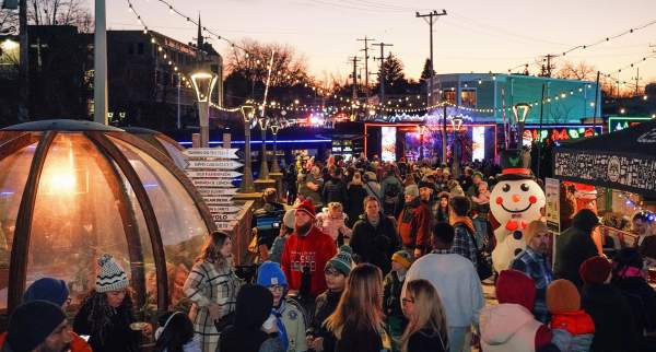 A large crowd attends an outdoor winter festival at dusk, with string lights, food tents, a clear dome shelter, and a large inflatable snowman