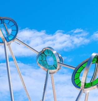 A modern metal and glass sculpture featuring three rings with green and blue glass accents is set against a bright blue sky and scattered white clouds.