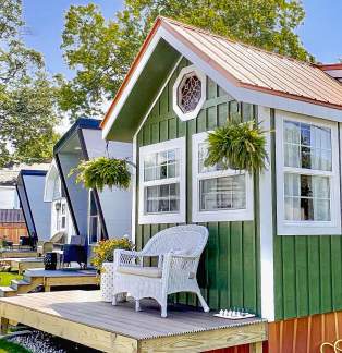 Green vertical-sided tiny house with a small front porch furnished with white wicker seating, set against a blue sky and leafy green tree.