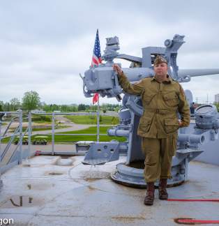 WWII reenactor in WW2 outfit stands before A three-inch 50 caliber naval gun on deck of USS LST 393 museum