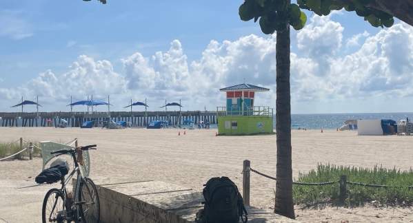 Bicycle standing at the edge of the beach with the Pompano Beach Pier and a lifeguard tower in the background.