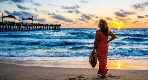 A woman standing at the ocean's edge watching the sunrise over the Pompano Beach Pier