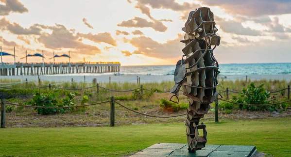 A sculpture of a seahorse sitting beachside on a patch of green grass with the Fisher Family Pier in the background at sunrise in Pompano Beach.