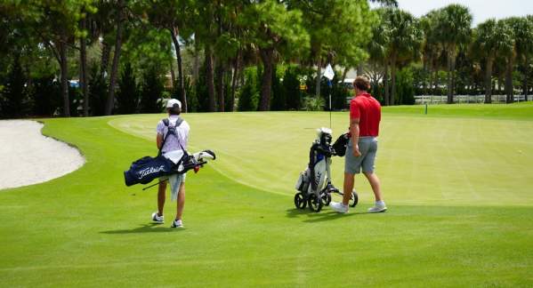 A father and son walking on a beautiful, green golf course in Pompano Beach. Dad's bag is on a push cart, and the son is carrying his bag.