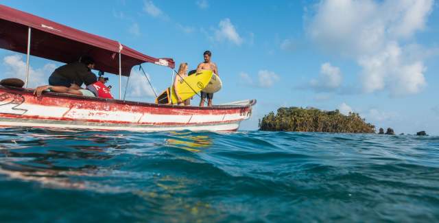 Group of people on a small boat, one holding a yellow surfboard. In the background, there are lush green islands and a clear blue sky.