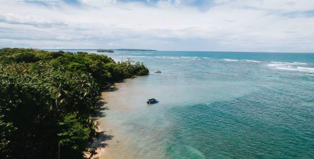 Sandy beach with a rocky shoreline, lush green vegetation, and a clear blue ocean with gentle waves. The sky is bright with a few fluffy clouds.