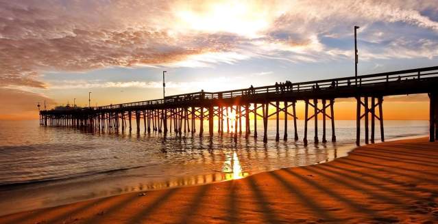 Newport Beach Pier at sunset with golden light reflecting on wet sand, long shadows stretching across the shoreline, and waves rolling in under a colorful sky.