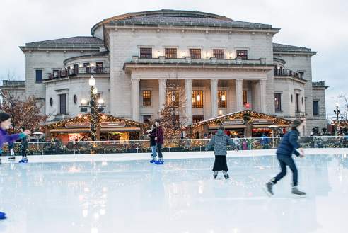 6 ice skaters on an ice rink in front of a building with holiday lights on it