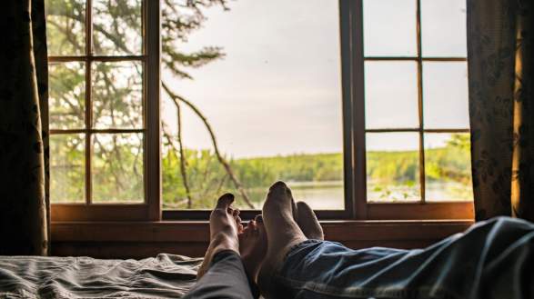 Two people's feet with the window showing view on the lake/wilderness from their cabin
