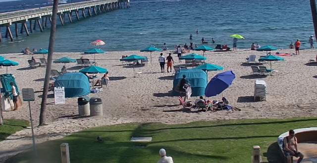 View of Deerfield Beach from the web cam at Wyndham Deerfield Beach. The pier is visible on the left side and people are lounging on beach chairs with umbrellas and cabanas