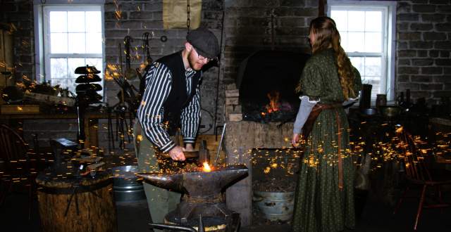 Blacksmith striking a hot iron on the anvil and making sparks fly. A fire and a female blacksmith in period dress are in the background