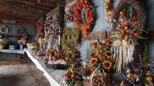 A rustic interior displays a variety of floral arrangements and wreaths on tables at the Bluemont Fair, showcasing vibrant colors and textures.
