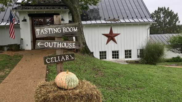 A rustic signpost points to The Barns at Hamilton Station Vineyard's, tasting room, outhouse, and cellar. A pumpkin rests on hay in the foreground.
