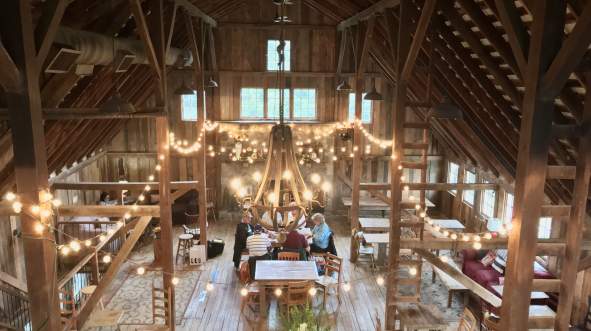 The side of The Barns at Hamilton Station Vineyards, a spacious, rustic barn interior with wooden beams, string lights, and a large chandelier. Guests gather around a long table, enjoying the ambiance.