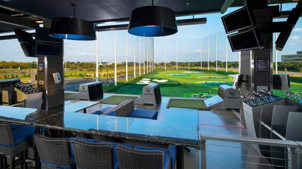 Top Golf driving range with a bar area, featuring high-tech screens and seating in Ashburn, Virginia. Lush green turf and targets visible outdoors.
