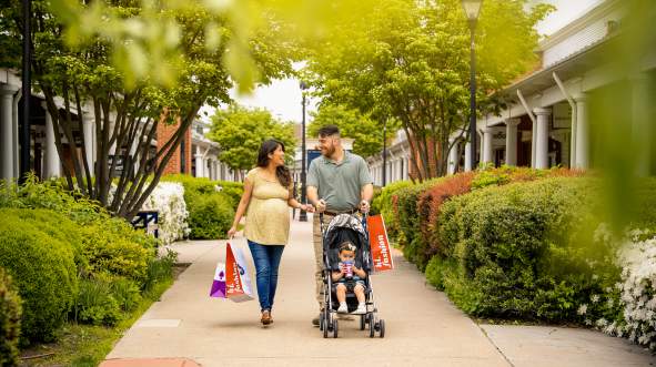 A couple strolls along inside Leesburg Premium Outlets, carrying shopping bags, with a child in a stroller. Lush greenery and shops line the walkway.