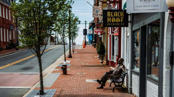 Bronze statue seated on a bench along the brick sidewalk of downtown Leesburg, with Black Hoof Brewing Co. and historic storefronts visible along King Street