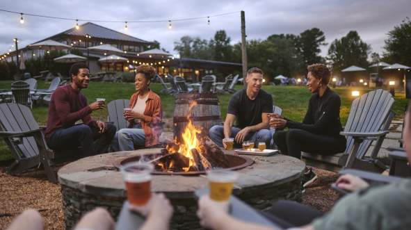 Group of friends enjoying craft beer at Lost Barrel Brewery around a firepit with scenic outdoor background and hanging lights at dusk