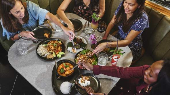 A group of four women share a variety of dishes at a round table, featuring vibrant plates of food and drinks at Sense of Thai in Ashburn, Virginia.
