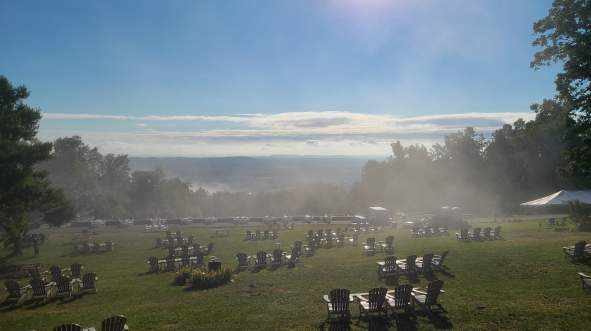 Bear Chase Brewing's misty landscape featuring numerous wooden chairs arranged on a grassy field, with a bright sun overhead and distant hills in the background.