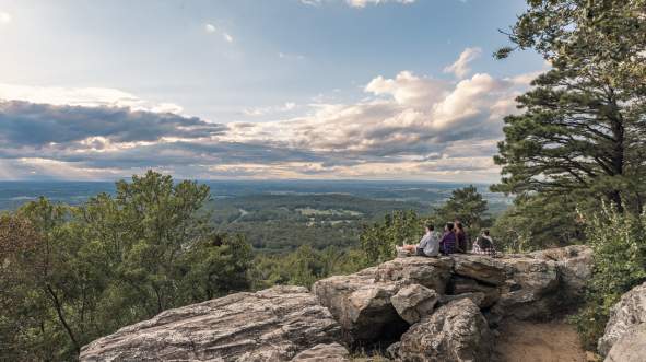 A group of five people sits on large rocks overlooking Bears Den Overlook in Bluemont, surrounded by trees and under a cloudy sky. The scene captures a serene landscape.