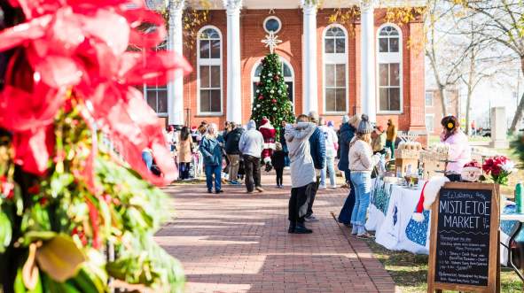 A festive outdoor market scene in front of Leesburg's historic Court House with a large Christmas tree, vendors selling goods, and visitors enjoying the atmosphere.