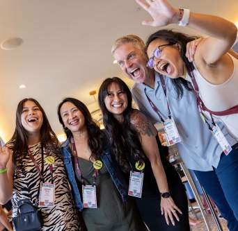 Group of 4 women and 1 man smiling and posing for the camera, angled up towards them from below.