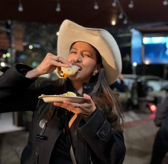 Woman in all black and a cowboy hat joyfully biting into a full taco.