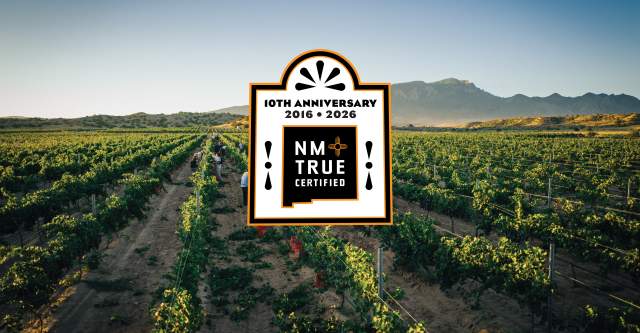 A vast vineyard stretches under a clear sky, with mountains in the background. In the center, there's a large "NM True Certified" logo celebrating its 10th anniversary from 2016 to 2026.