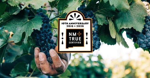 A hand carefully holds a bunch of ripe grapes on a vine. The image features a "NM True Certified" badge, celebrating its 10th anniversary from 2016 to 2026.