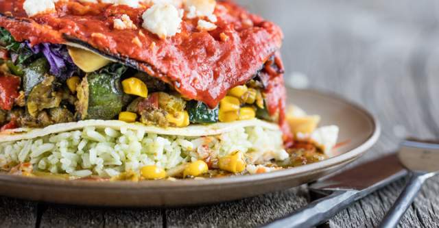 a colorful stack of different types of ingredients stacked between corn tortillas sit on a plate, next to silverware on a wooden table