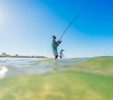 A man is fishing from the shore as his son plays in the waters beside him.