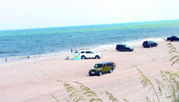 Off-road vehicles parked on Outer Banks sandy beach with visitors walking along the shore and turquoise water in bright sunlight.