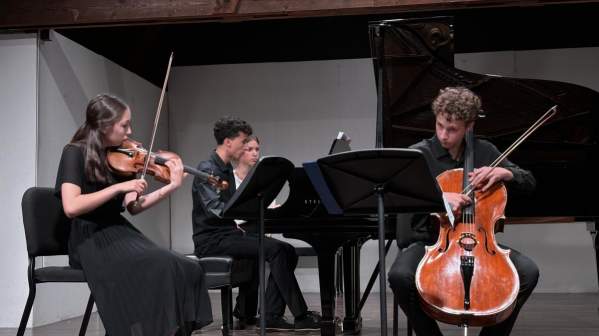 Four Luzerne Music Center students playing violin, cello and piano on stage indoors with white wall in back