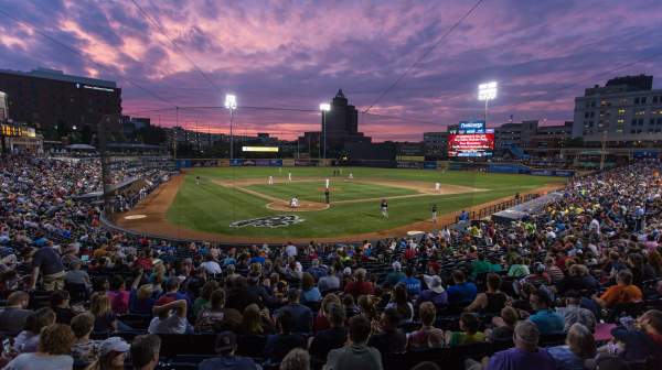Crowded Akron Rubber Ducks baseball stadium at sunset with players on the field and a vibrant purple-pink sky overhead.