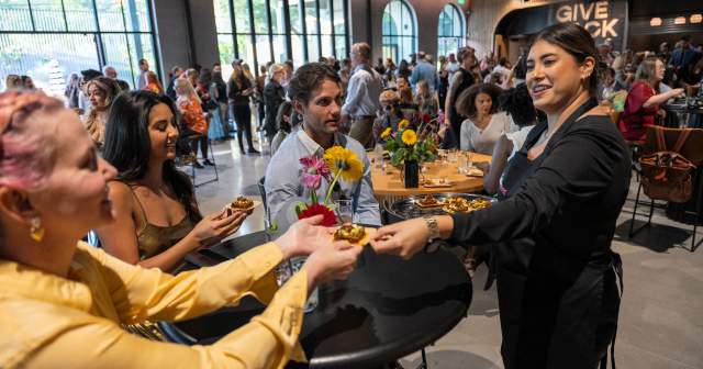 Student chef handing a bite of food to someone at a table of Give Back Gig attendees.