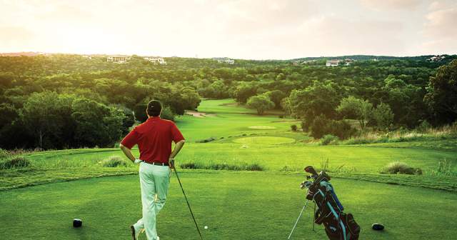 Male golfer stands on a teebox with driver and bag looking out towards a sunset