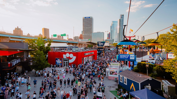A large crowd moves through the bustling Summerfest grounds in Milwaukee during the day, with the downtown skyline visible in the background. A red building with the Summerfest smiley-face logo serves as the festival store. Above, colorful gondolas on the Skyglider transport people across the venue. The scene is filled with booths, food stands, and signage under a clear sky.
