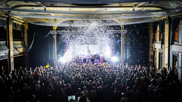 Interior view of Turner Hall ballroom during a live concert, showing a packed crowd of hundreds of people facing an illuminated stage. The historic venue's ornate architecture is visible with decorative columns, balconies, and detailed ceiling work. Dramatic stage lighting creates bright white beams cutting through the dark space, with confetti or snow effects falling from above.