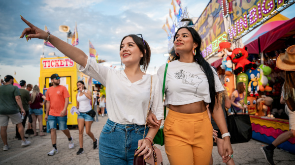 Two young women walk arm-in-arm through the Wisconsin State Fair, with one pointing excitedly ahead. Carnival rides, game booths, and crowds create a festive atmosphere in the background.