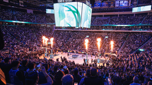 A packed crowd fills Fiserv Forum in Milwaukee during an NBA game as flames shoot up from the court during player introductions, with the arena’s large video scoreboard displaying a close-up of a player.
