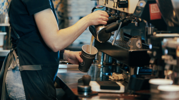 A barista pours steamed milk into a paper cup at an espresso machine inside a coffee shop.