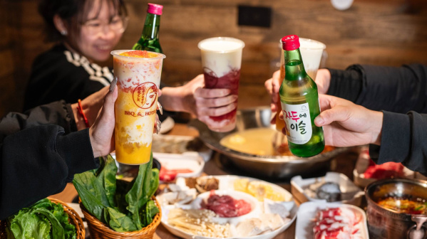 Friends raise colorful drinks and a bottle of soju over a table filled with Korean BBQ and hot pot, celebrating a lively, shared dining experience.