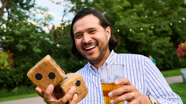 Man holding large wooden dice and a beer at a beer garden.