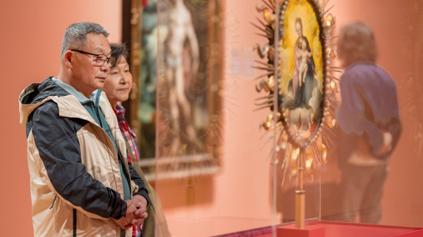 A middle-aged man wearing glasses and a tan-and-black jacket stands with his hands clasped, looking thoughtfully at an artwork in what appears to be an art museum or gallery. Behind him, a woman stands slightly out of focus, also observing the exhibit.

In the foreground on the right, there is a blurred religious-style artwork or icon displayed in a glass case, with a gold decorative frame. The overall scene has warm lighting and a calm, contemplative mood.
