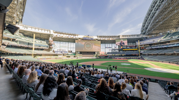 Crowd of business attendees seated in American Family Field's lower bowl during the Visit Milwaukee annual meeting, with the baseball stadium's retractable roof open and a presentation screen visible on the outfield wall.