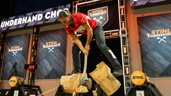 A STIHL TIMBERSPORTS athlete competes in the underhand chop event, standing on a log and swinging an axe to split the wood. Wood chips fly across the stage as competition signage and score displays frame the high-intensity lumberjack championship.