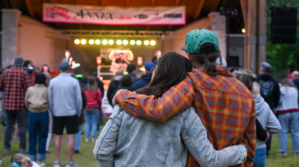 A couple stands arm in arm watching a live outdoor concert from the audience. Stage lights glow beneath a covered pavilion as community members gather on the lawn to enjoy the performance.