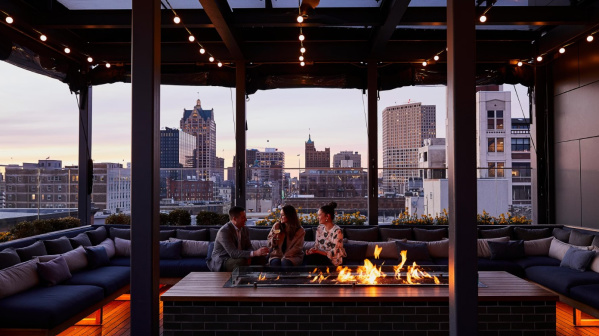Three people sitting around a rooftop fire pit at sunset, enjoying drinks with a view of downtown Milwaukee’s skyline at The Outsider rooftop bar.